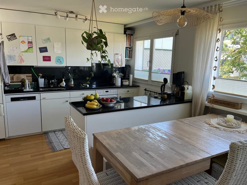 Modern kitchen with white cabinets, black countertops, and a wooden dining table with wicker chairs. Features include a pendant light, a hanging plant, and various kitchen appliances.