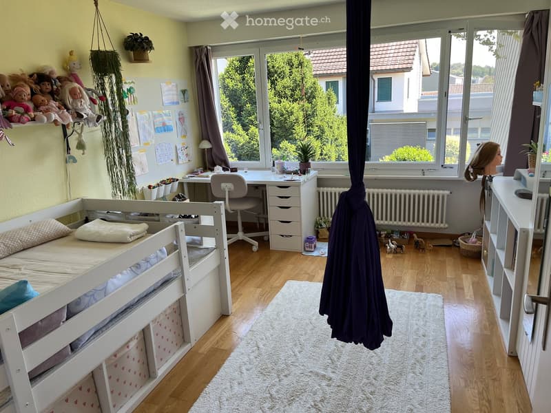 Bright bedroom with a white bunk bed, desk, chair, shelves, and toys. Large windows offer a view of greenery outside.