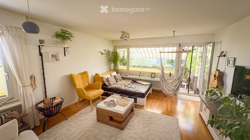Bright living room with wooden flooring, large windows, a yellow armchair, a sofa with pillows, a coffee table, a hammock, and a guitar on the wall.