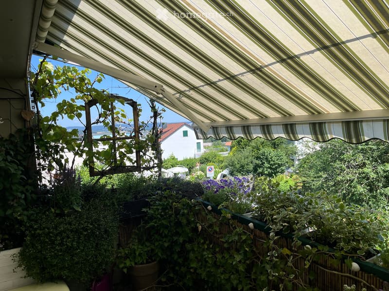 Balcony with striped awning, lush greenery, and a scenic view of the sea and town.