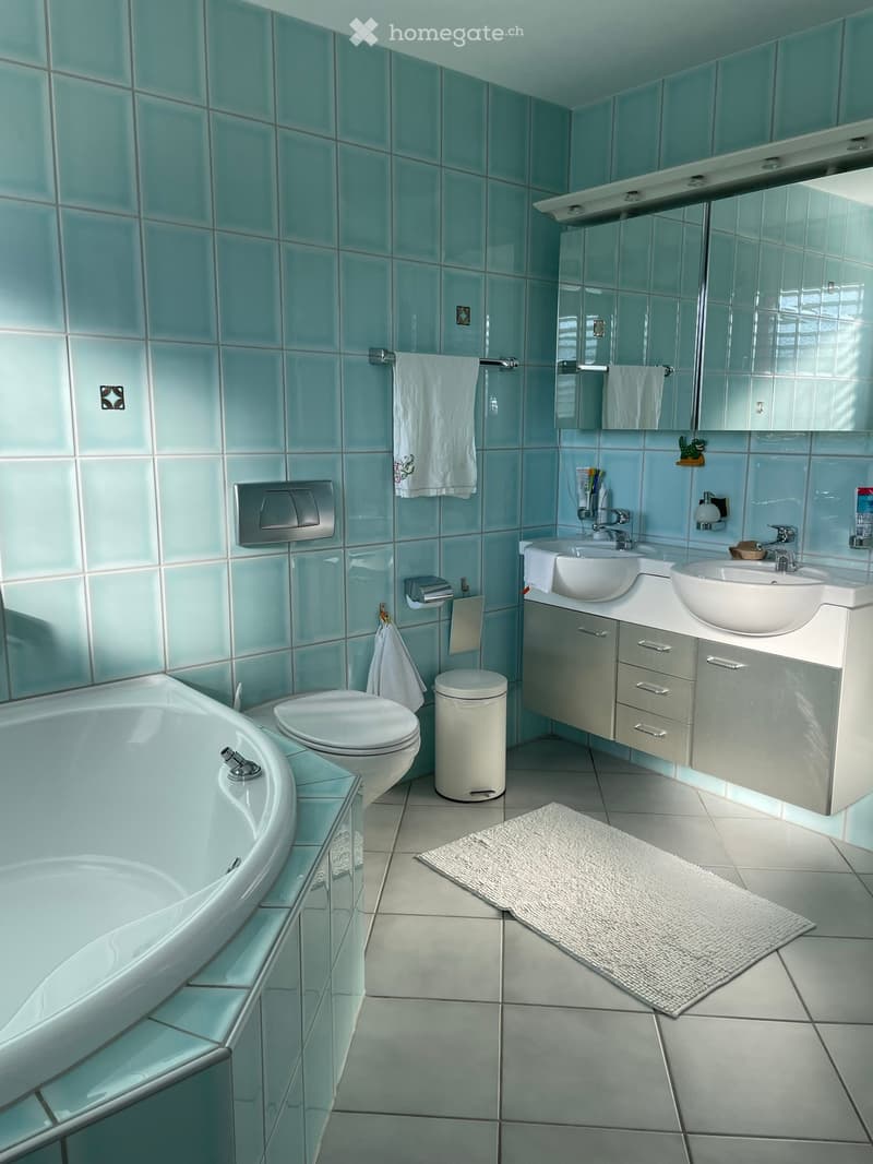 Modern bathroom with light blue tiles, white fixtures, double sink vanity, bathtub, toilet, and a small trash can. Includes a white towel and a bathroom rug.
