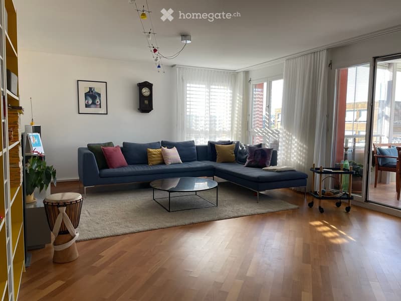 Modern living room with a blue sectional sofa, colorful throw pillows, a glass coffee table, a drum, a small table with wheels, and large windows with sheer curtains. The room features wooden flooring and a white rug.
