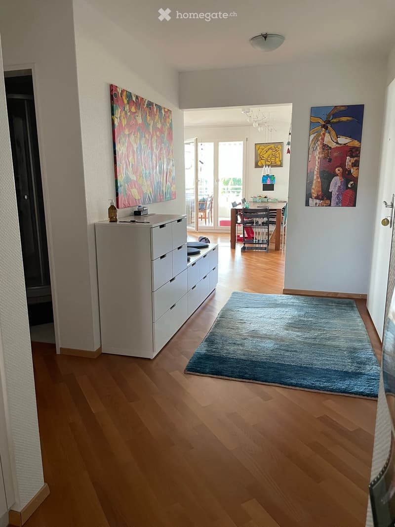 Hallway with wooden flooring, white cabinet, blue rug, and artwork on walls. Open doorway leads to a dining area with a table and chairs.