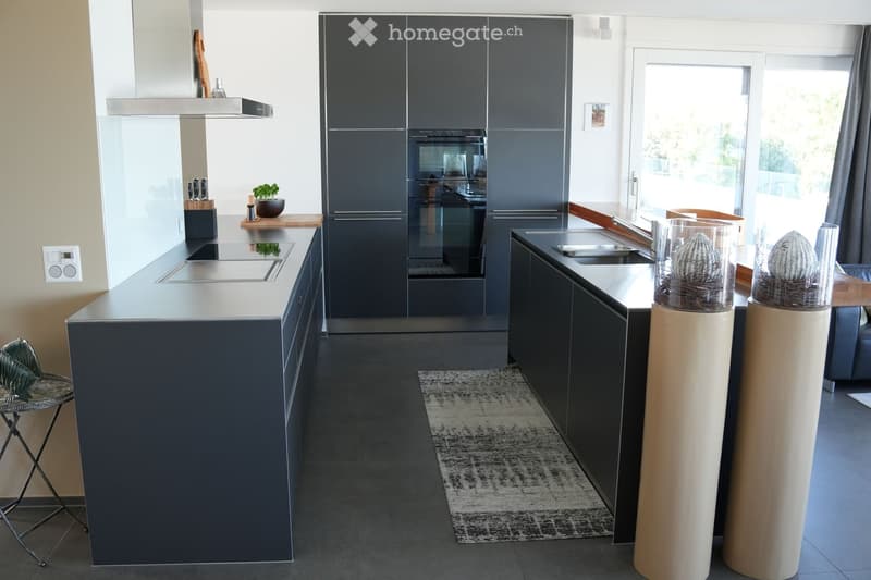 Modern kitchen with dark grey cabinets, stainless steel appliances, island with integrated stove, sink, and seating area. Features a small potted plant, knife block, and decorative columns.