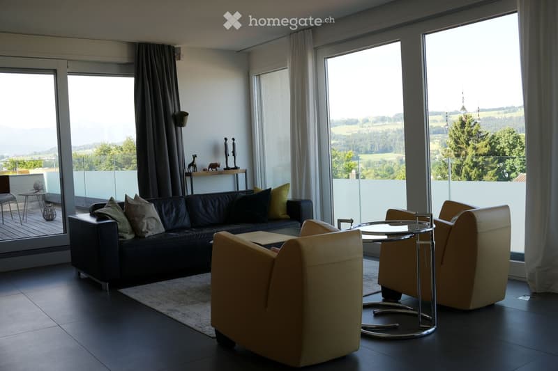 Modern living room with large windows offering a scenic view, featuring a black leather sofa, cream armchairs, a round glass coffee table, and a small side table with decorative items.