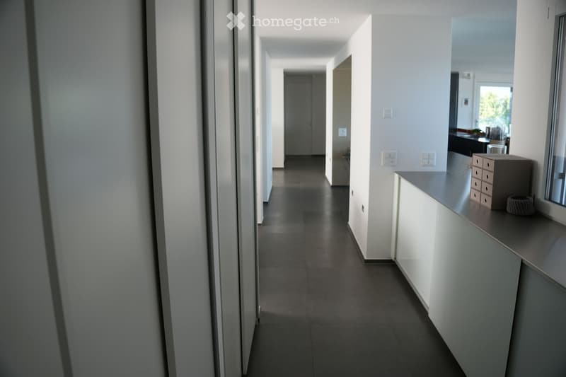 Modern hallway with white walls, dark tiled floor, and recessed lighting. Features a small console table with a mirror, decorative items, and a glimpse of a room with furniture in the background.