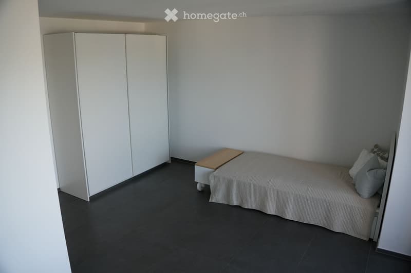 Modern, minimalist bedroom with a single bed, white wardrobe, and a small nightstand. The room features dark tiled flooring and light-colored walls.
