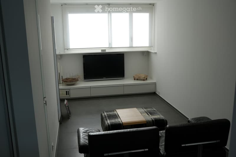 Modern living room with dark tiled flooring, white walls, and large windows. Features a sofa, coffee table, television on a white cabinet, and decorative items including a bowl and candles.