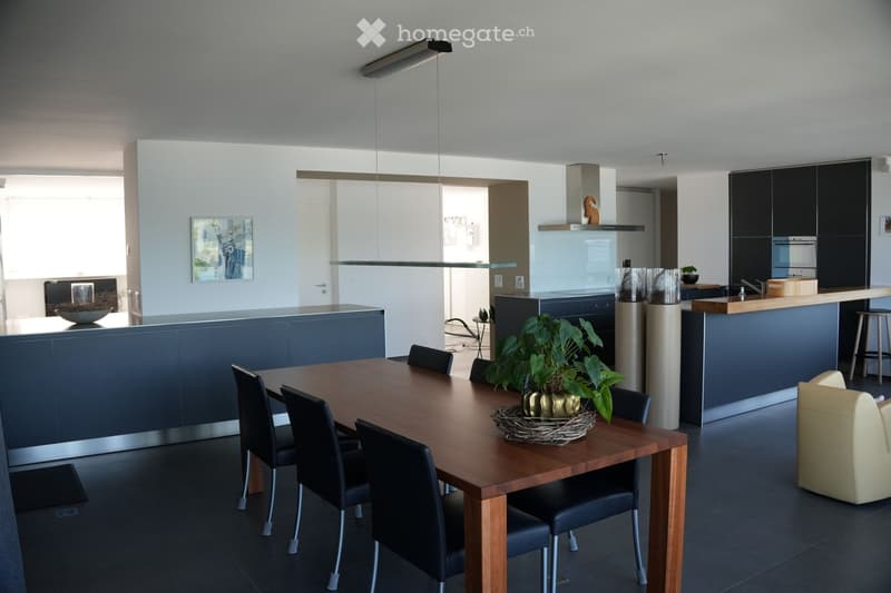 Modern open-plan kitchen and dining area with dark blue cabinetry, a wooden dining table with black chairs, a kitchen island, and a small seating area with a cream-colored armchair.