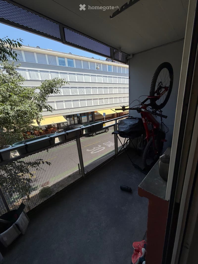 Balcony with a bicycle, railing, and view of a building and outdoor seating area.