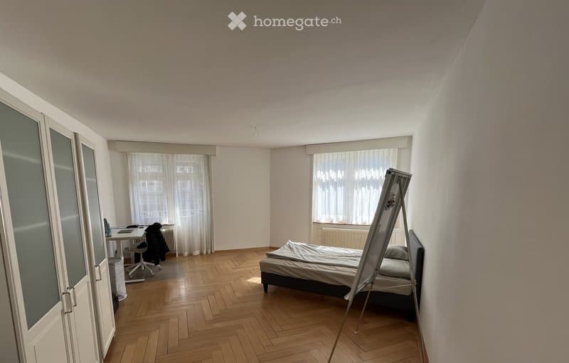 A furnished bedroom with a wooden floor, a bed, a desk with a chair, and a wardrobe with mirrored doors. The room has large windows with white curtains.