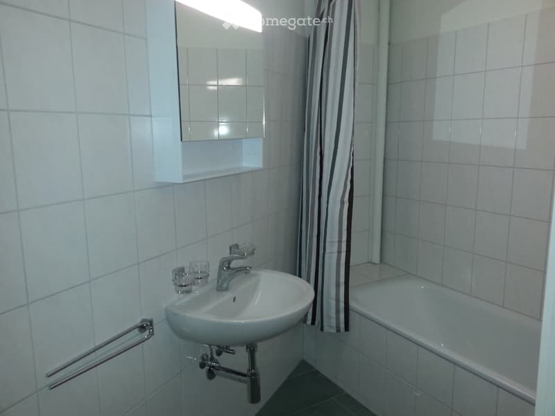 A small bathroom with a white tiled wall, a sink with a faucet, a bathtub with a striped shower curtain, a mirror with a built-in shelf, and a handrail for support.