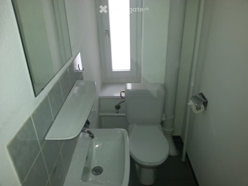 A small bathroom with a white sink, toilet, bathtub, and a mirror above the sink. The walls are tiled, and there is a window allowing natural light to enter.