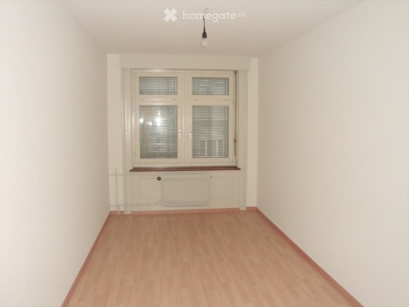 Empty room with light wooden flooring, white walls, and a large window with blinds. A radiator is visible below the window, and a single light fixture hangs from the ceiling.
