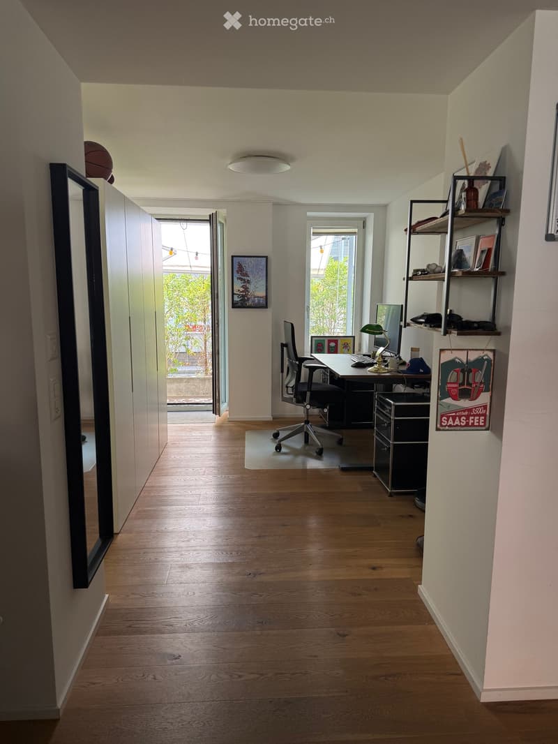 Office space with wooden flooring, large windows, desk, chair, computer, lamp, shelves, and artwork.