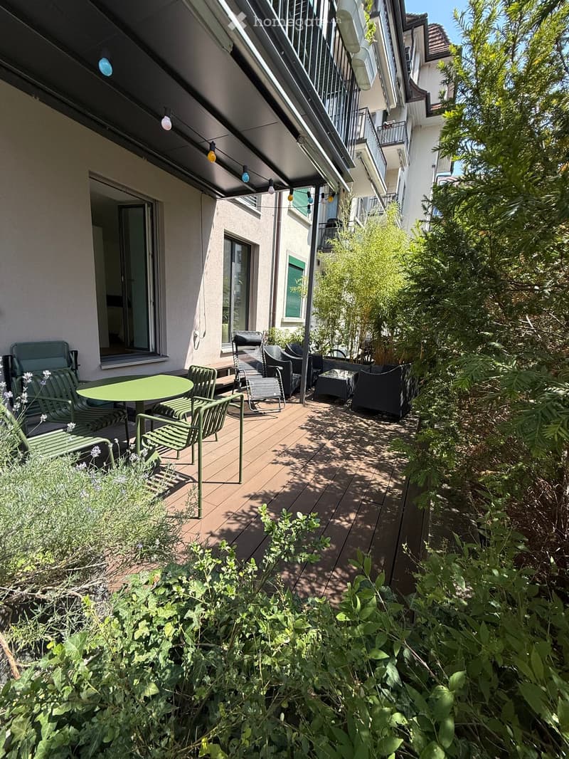 Balcony with wooden decking, green table, and several chairs, surrounded by lush greenery and plants.