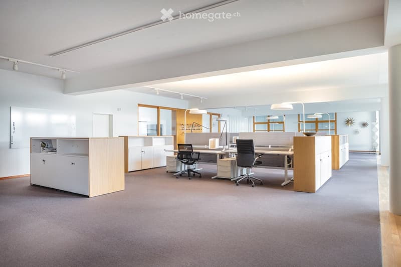 Modern office space with desks, chairs, cabinets, and a large screen on the wall. The room features a gray carpet, white walls, and large windows with wooden frames.
