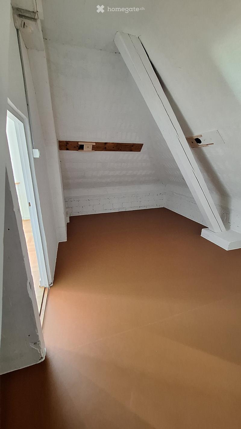 Empty attic space with exposed wooden beams, unfinished walls, and a slanted ceiling. The floor is painted brown, and there is a partially open door leading to another area.