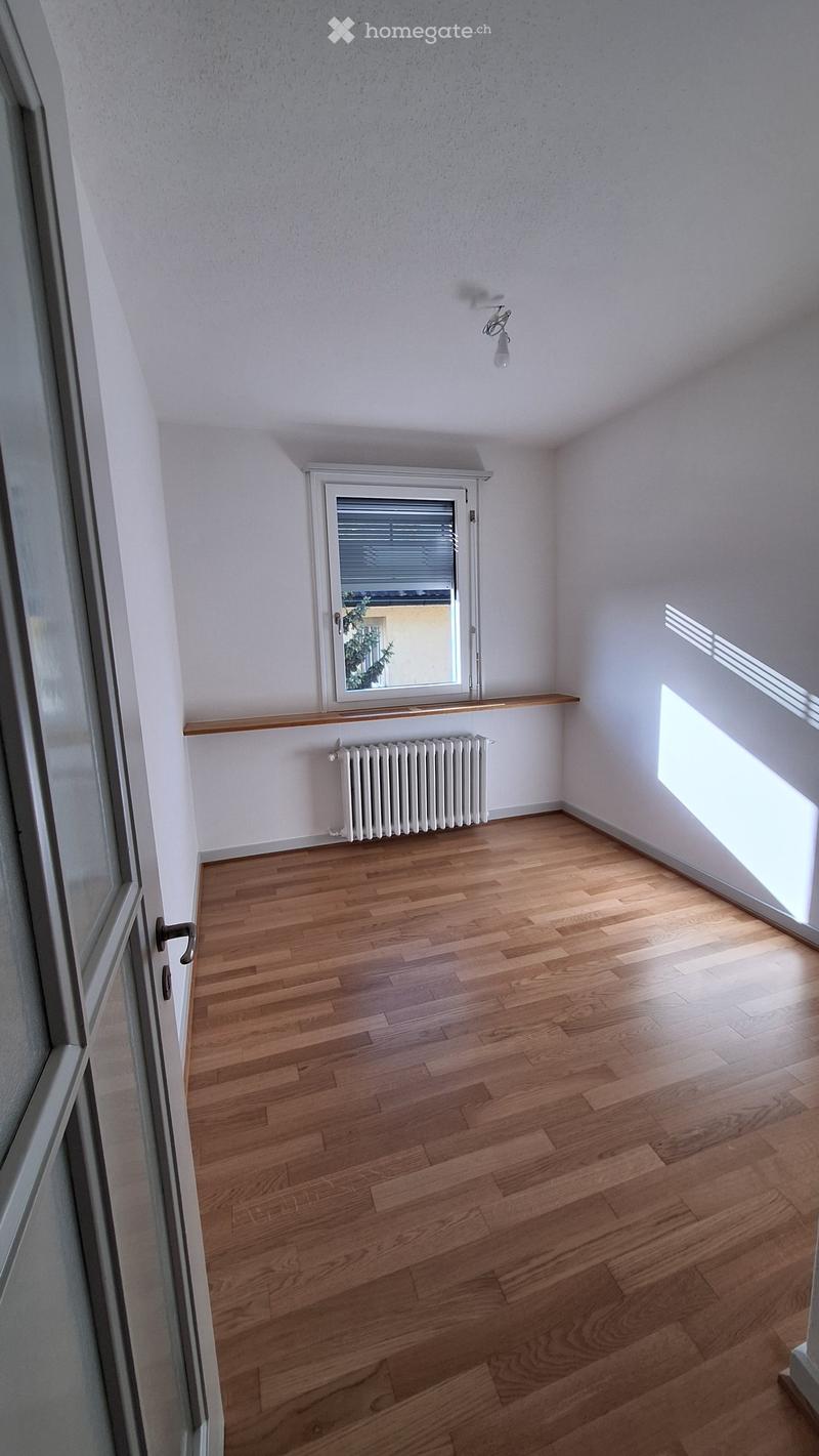 Empty room with wooden flooring, white walls, a window with blinds, a radiator, and a small wooden shelf above it.