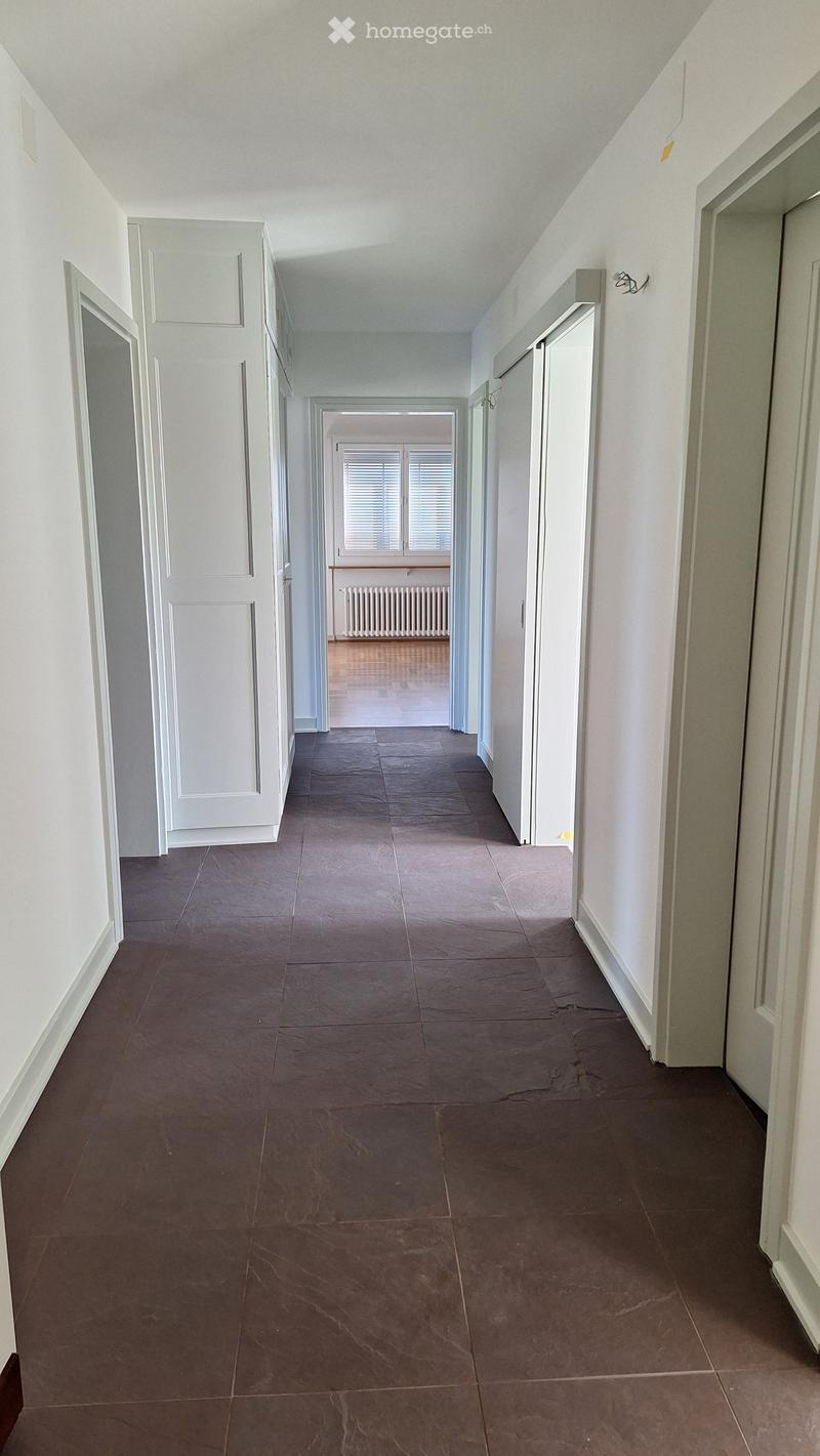Empty hallway with brown tiled floor, white walls, and multiple doors on both sides. Natural light comes from a window at the end of the corridor.