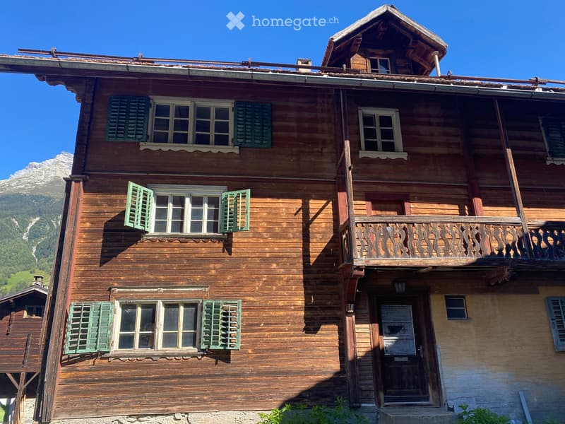 A two-story wooden house with green shutters, a balcony, and a small attic window, set against a mountain backdrop.