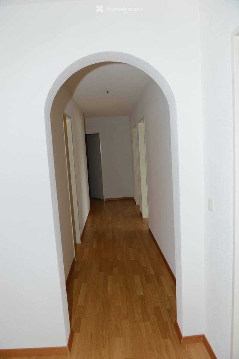 A long, narrow hallway with wooden flooring and white walls, featuring an arched ceiling and several doors along the sides.