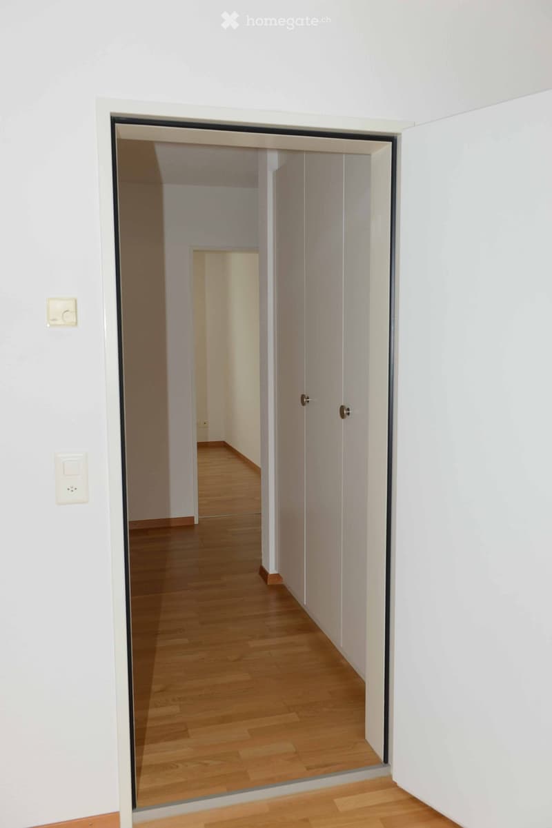 Empty hallway with light wooden flooring and white walls, featuring a partially open door leading to another room.