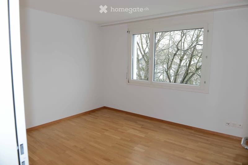 Empty room with light wooden flooring and white walls, featuring a large window with white frame showing bare trees outside.