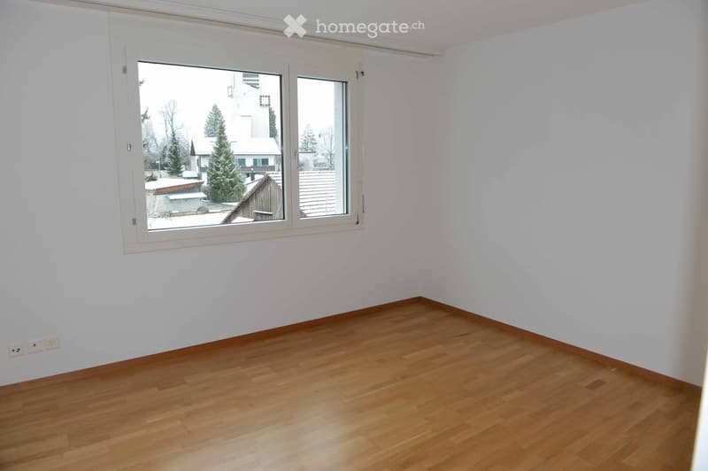 Empty room with light wooden flooring and white walls, featuring a large window with white frame offering a view of the outside.