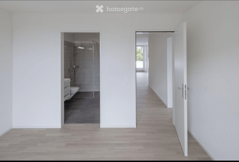 Bright, minimalist hallway with light wooden flooring and white walls. Open doors lead to a bathroom and another room.