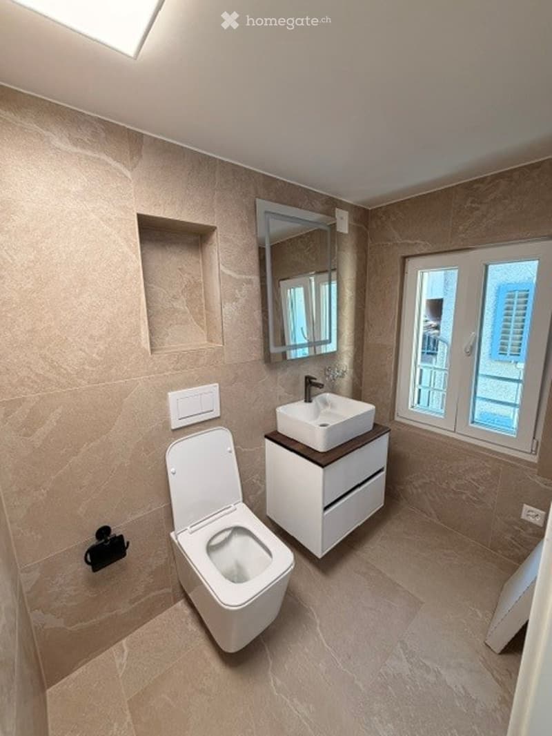 Modern bathroom with beige tiled walls and floor, white toilet, white sink with cabinet, mirror above sink, and a window providing natural light.