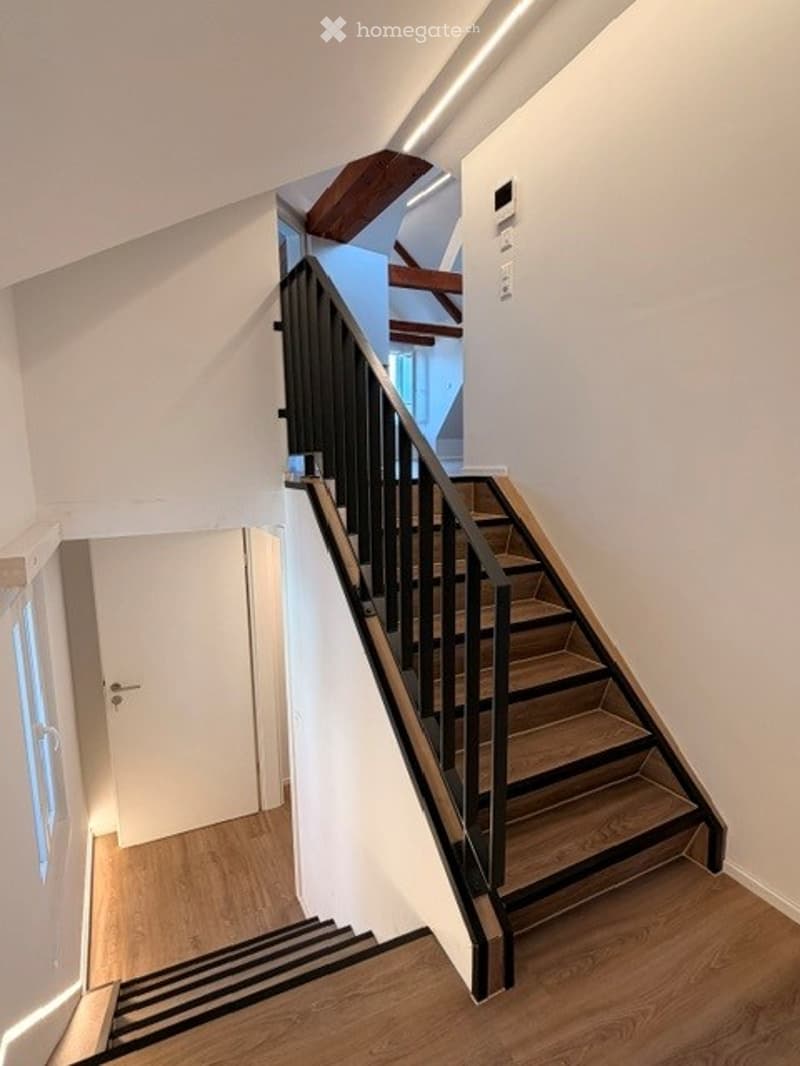 Modern interior staircase with wooden steps and black metal railing, leading to an upper level with exposed wooden beams.