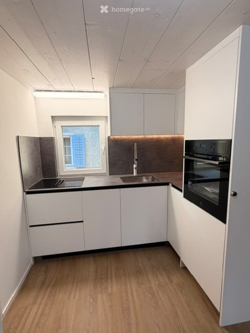 Modern, minimalist kitchen with white cabinets, a black countertop, a sink with a faucet, and an oven. The space features light wooden flooring and a window offering a view of a building outside.