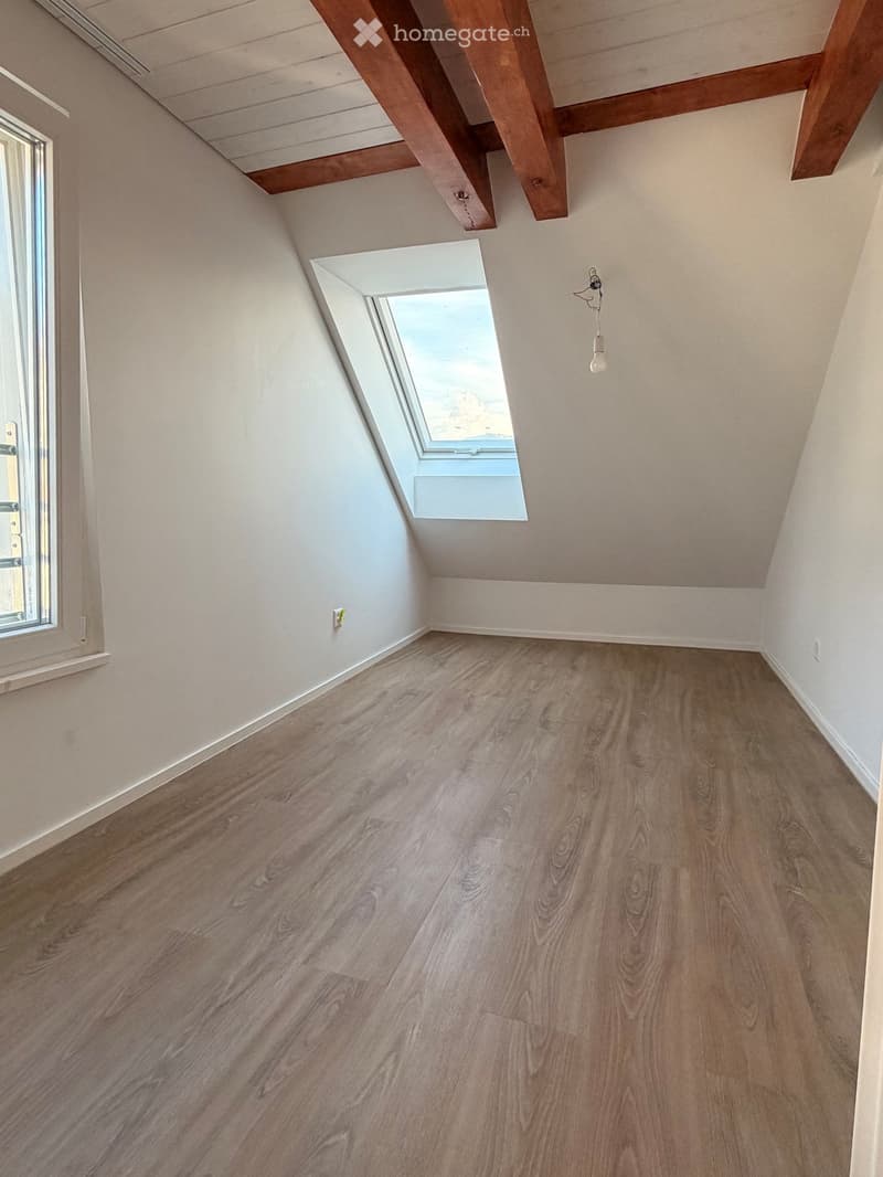 Empty attic room with wooden flooring, white walls, exposed wooden beams, and a skylight window offering a view of the sky.