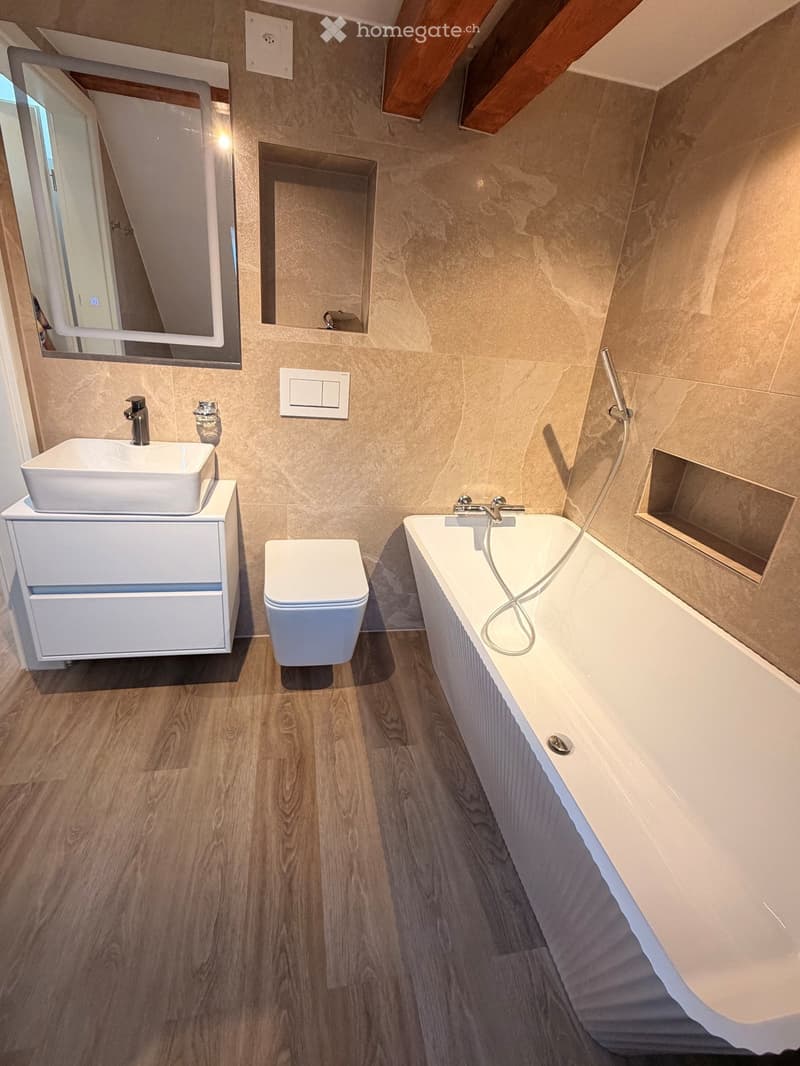 Modern bathroom with wooden flooring, white sink, toilet, bathtub, and large mirror. Exposed wooden beams on the ceiling.
