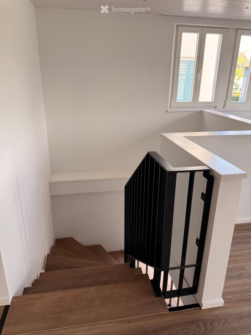 Staircase with wooden steps and a black metal railing, white walls, and a window at the top landing.