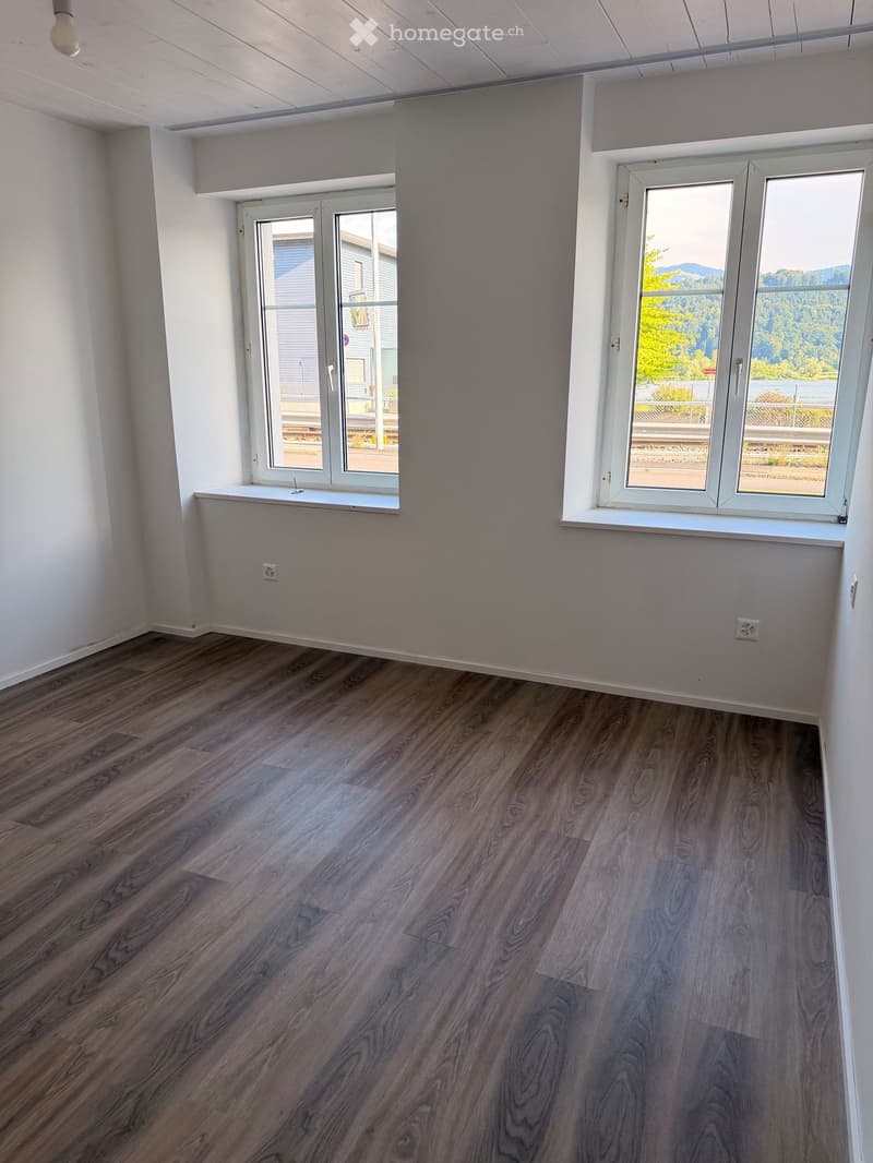 Empty room with wooden flooring, white walls, and large windows offering a scenic view of trees and a road.