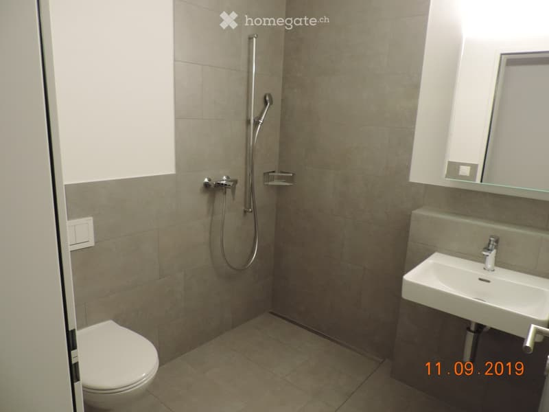 Modern bathroom with beige tiled walls and floor, featuring a toilet, a sink with a mirror, and a shower with a handheld sprayer.