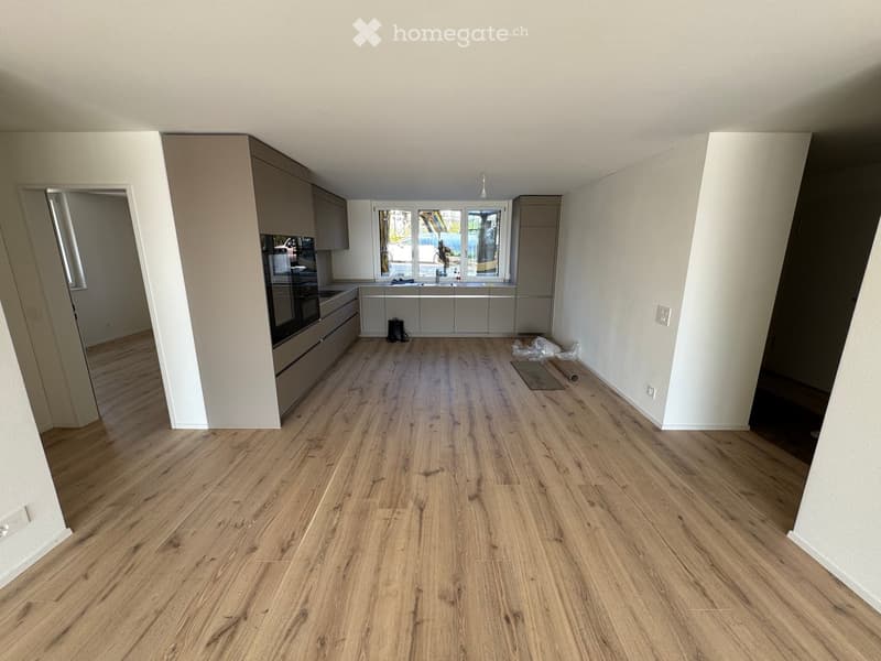 Empty kitchen with light wooden flooring, white walls, and fitted cabinets. The space includes a window with a view of the outside, and the room is currently unfurnished.