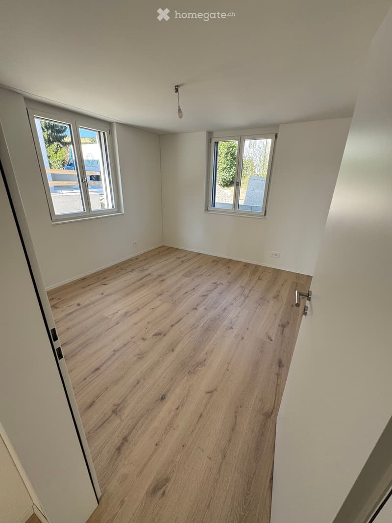 Empty room with light wooden flooring, white walls, and two large windows with metal frames. The room has a simple, clean design with no furniture or decorations.