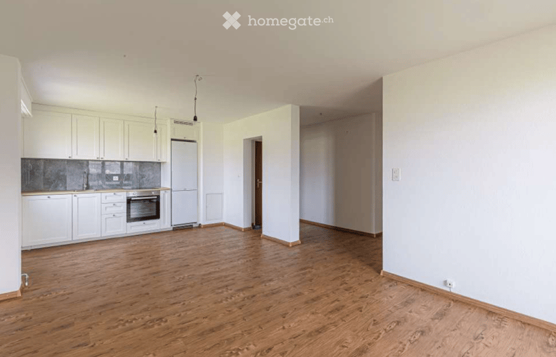 Empty room with wooden flooring, white walls, and a small kitchen area featuring white cabinets, a sink, and a refrigerator.