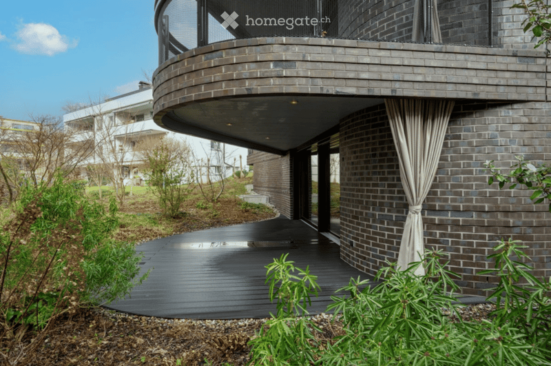 Modern two-story building with brick facade, curved balcony, and wooden deck surrounded by greenery.