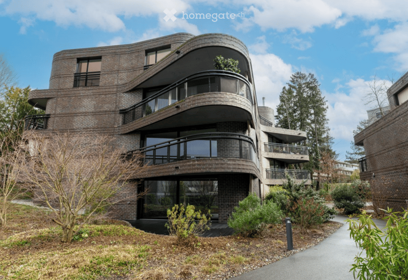 Modern, curved brick building with multiple levels, large windows, and balconies overlooking a landscaped garden area.