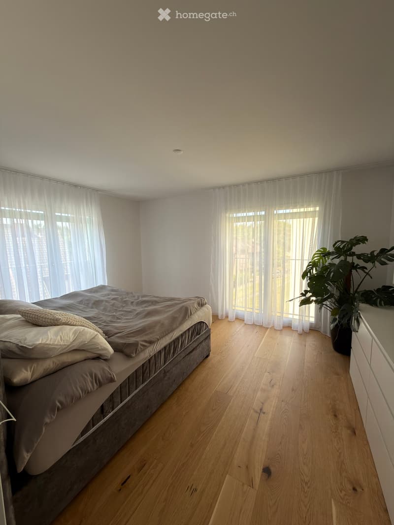 Modern bedroom with wooden flooring, large windows with sheer curtains, a neatly made bed with pillows and a blanket, and a potted plant near the window.