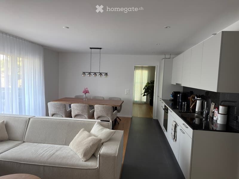 Open-plan living and dining area with a grey sofa, coffee table, dining table with chairs, and a modern kitchen with white cabinets, stainless steel appliances, and a sink.