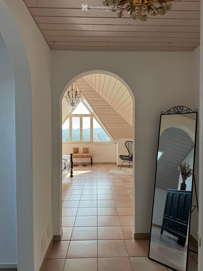 A spacious hallway with tiled flooring, light wooden ceiling, and an arched opening leading to a sunlit room with a bench, chair, and view of nature.
