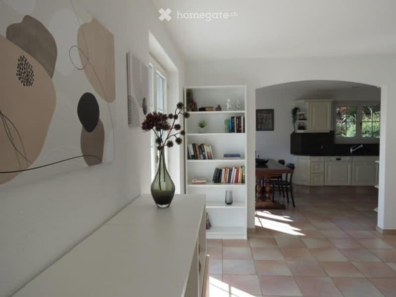Bright living room with white walls, large windows, a white bookshelf filled with books, a vase with flowers on a table, and an open view into a modern kitchen area with cabinets and a dining table.