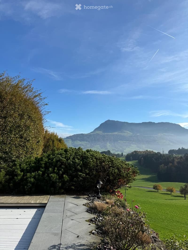 A serene garden area with a paved pathway, wooden deck, and lush greenery, offering a panoramic view of a mountainous landscape under a clear blue sky.