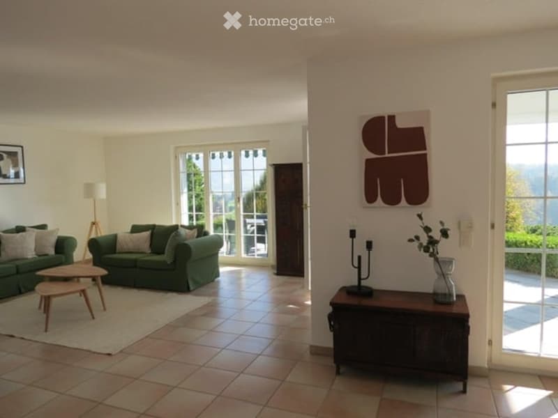 Bright living room with large windows offering a scenic view, green sofas, wooden coffee tables, a floor lamp, and a decorative table with a vase.