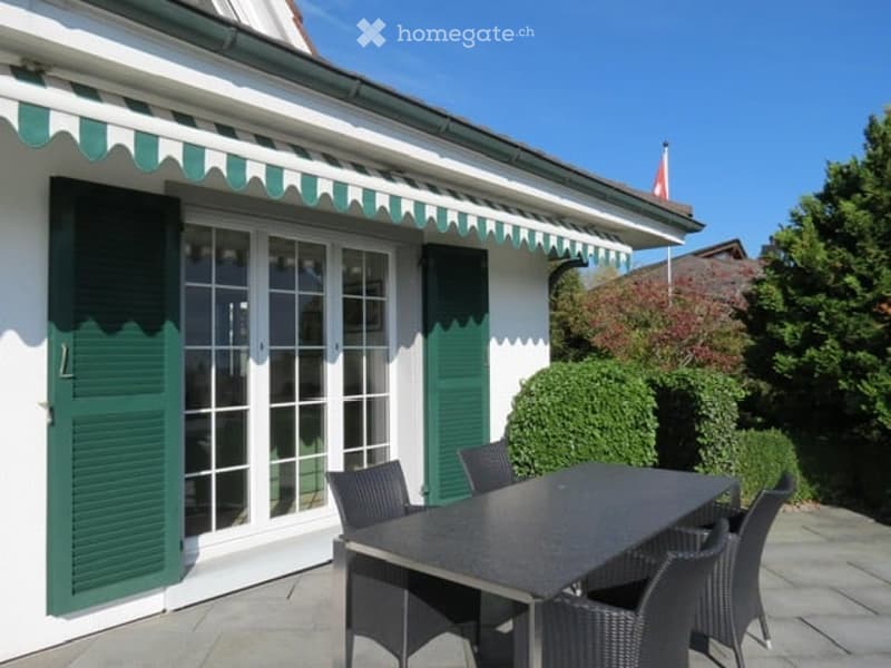 Balcony with a black dining table and four black chairs, green shutters, and a striped awning above.